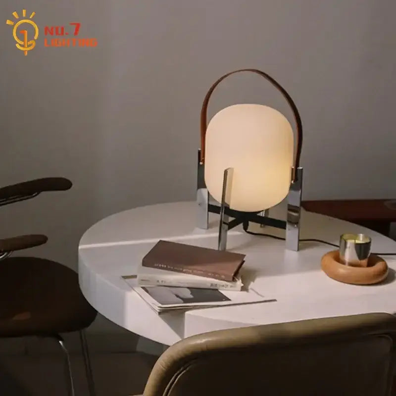 Modern cream designer metal glass table lamp with warm oval shade, soft glow, chrome base, and brown leather handle for bedroom decorbites