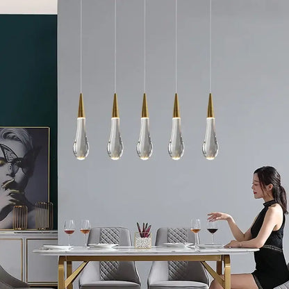 Five decorbites crystal gold pendant lights with clear teardrop glass shades hang above a dining table
