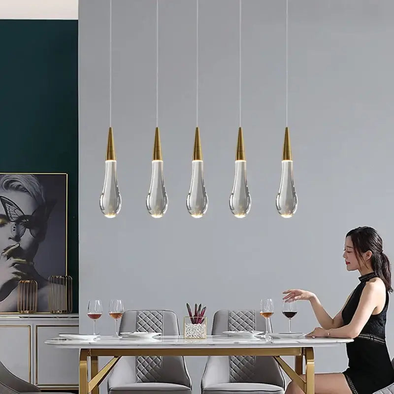 Five decorbites crystal gold pendant lights with clear teardrop glass shades hang above a dining table