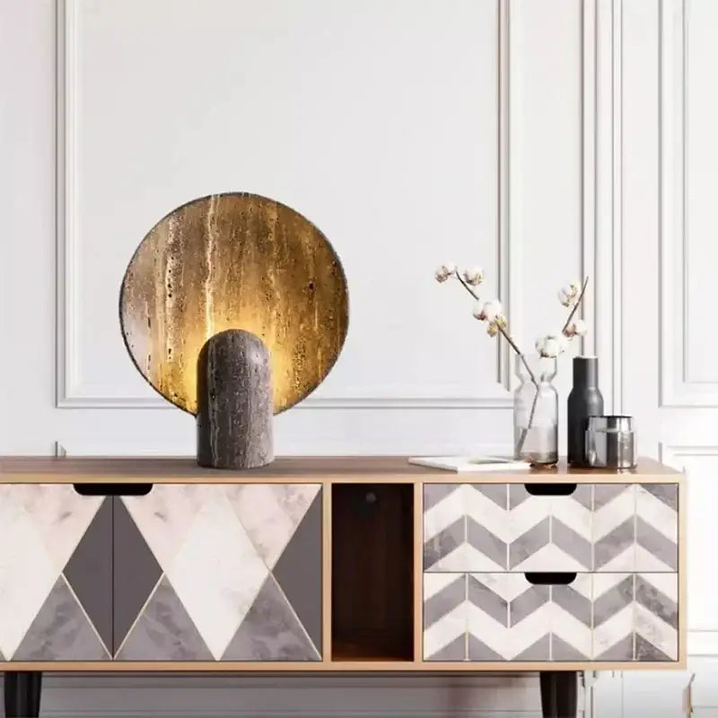 Modern circular table lamp with textured gold disc and dark travertine rock base for warm bedroom glow