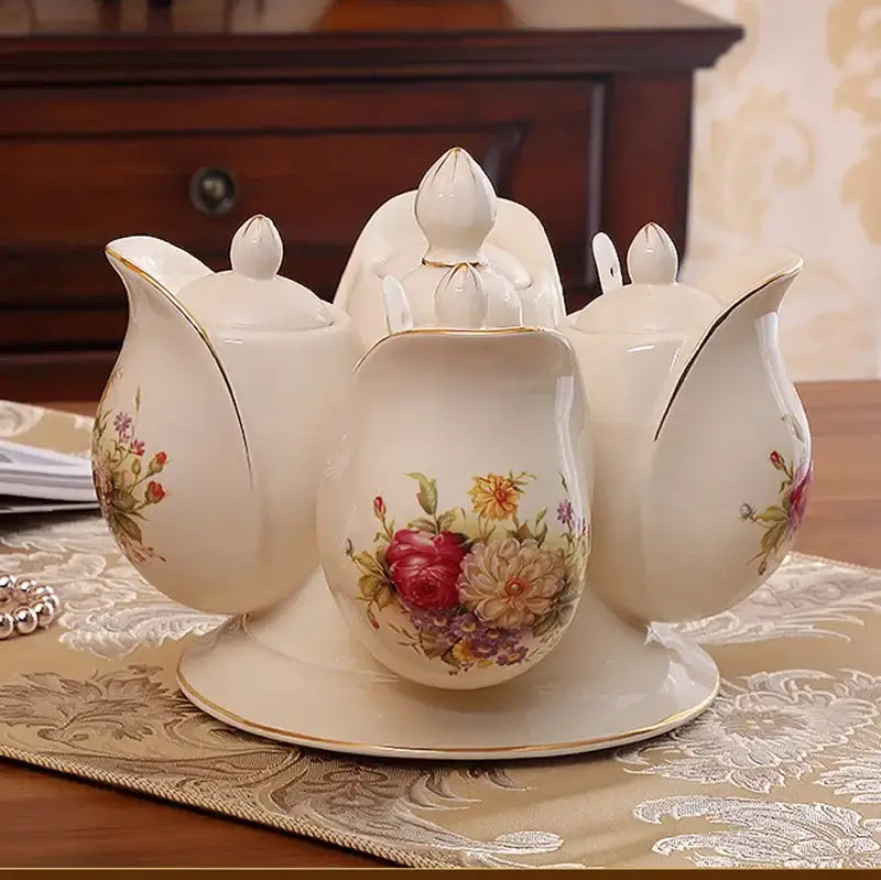 Cream-colored porcelain tea set with floral hand-painted designs and gold-trimmed lids beside deep green rotatable spice jar seasoning set