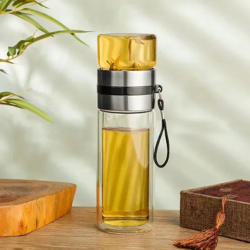 Clear double-walled glass tea infuser bottle with silver lid and black strap filled with golden tea for kitchen use