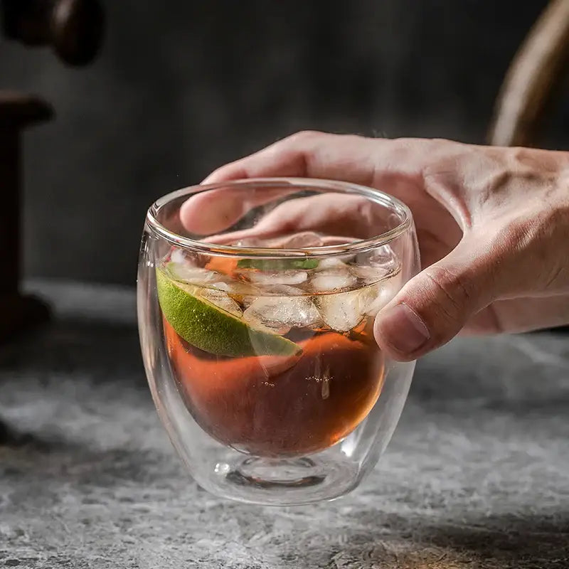 Modern double walled transparent glass cup holding amber liquid with ice and lime wedge in kitchen and dining room use