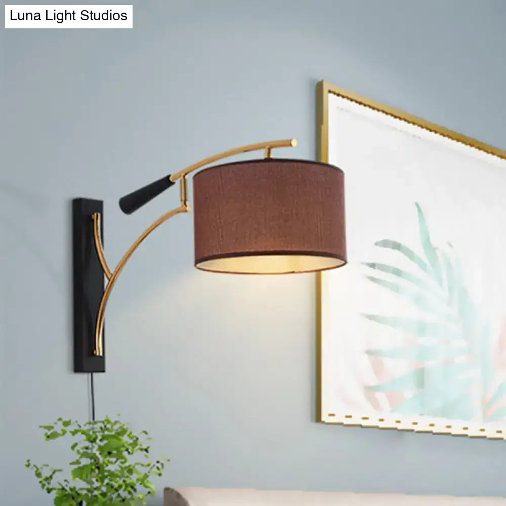 Modern drum shade wall lamp with a curved gold-toned arm, flaxen coffee fabric shade, and black handle grip for bedroom