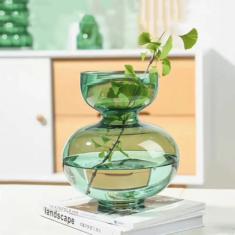 Modern emerald green gourd vase made of translucent teal glass holds delicate green ginkgo leaves in living room terrarium