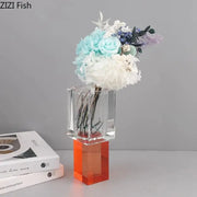Clear acrylic geometric square vase with vibrant orange base holding artificial flowers in blue white and purple hues decorbites geometric square decor