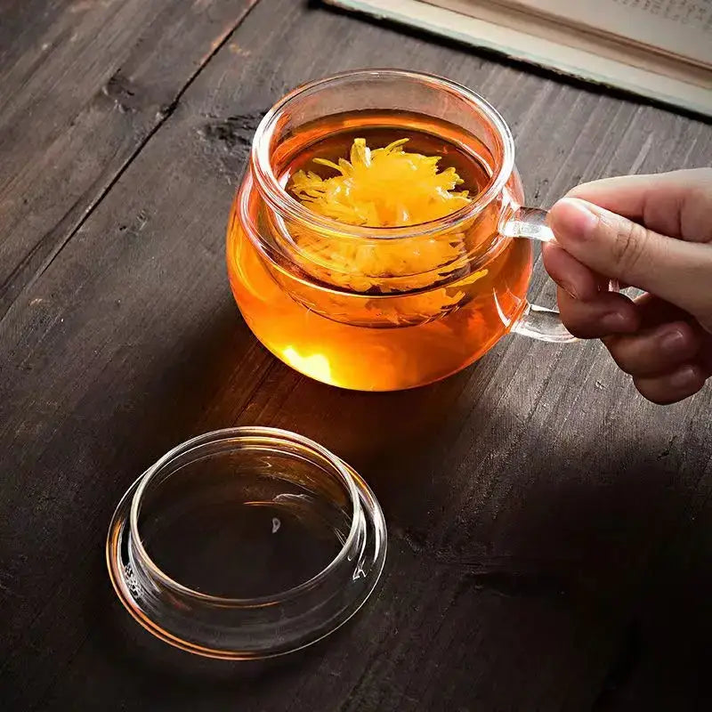 A modern clear glass tea cup with infuser and lid filled with amber glass tea and floating yellow flower petals