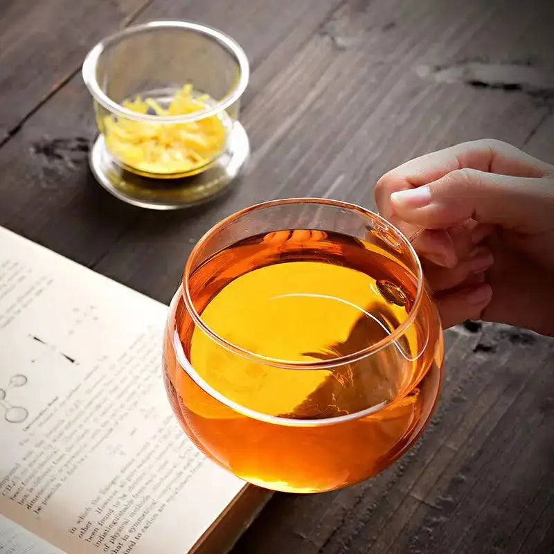 A hand holds a modern clear glass tea cup with infuser and lid filled with amber-colored glass tea
