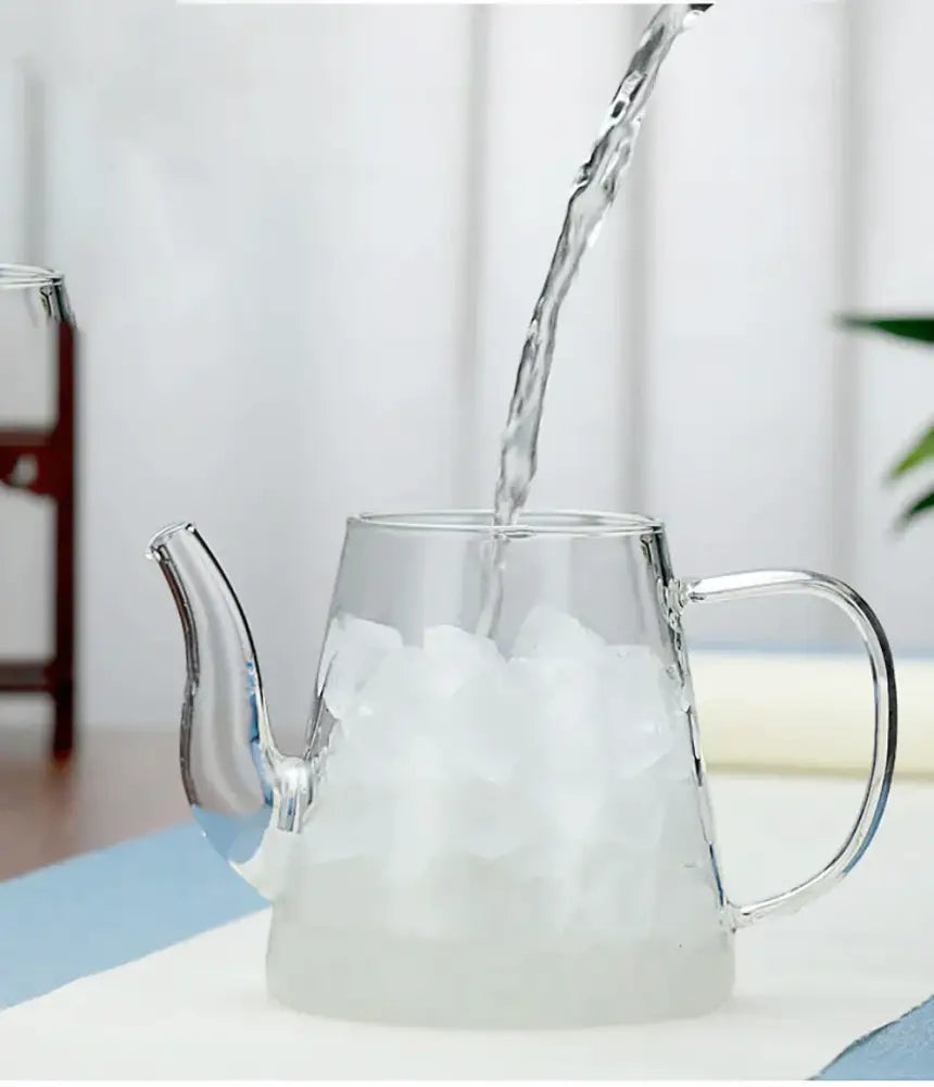 Clear glass tea infuser pot with curved spout and handle filled with ice cubes and water pouring in