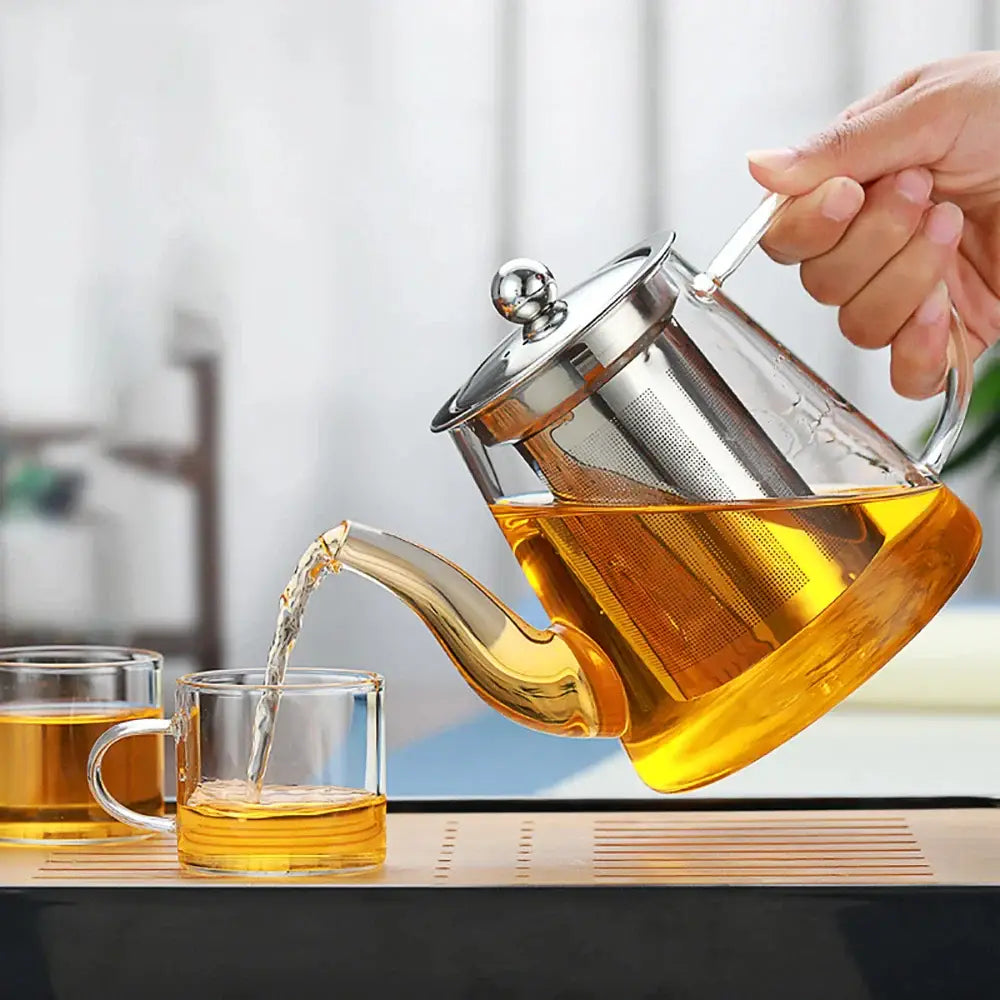 Clear glass tea infuser pot with stainless steel lid and built-in filter pouring tea into matching glass cups