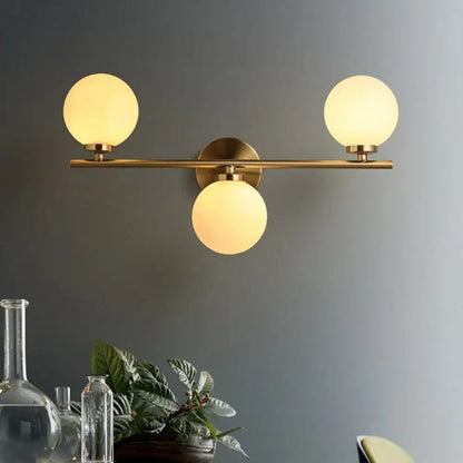 Brass-finished modern wall light with three white glass shades for bedroom featuring frosted glass globes