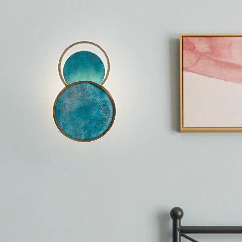 Modern wall sconce featuring two overlapping circular glass panels with a textured, teal-blue finish encased in polished brass frames.