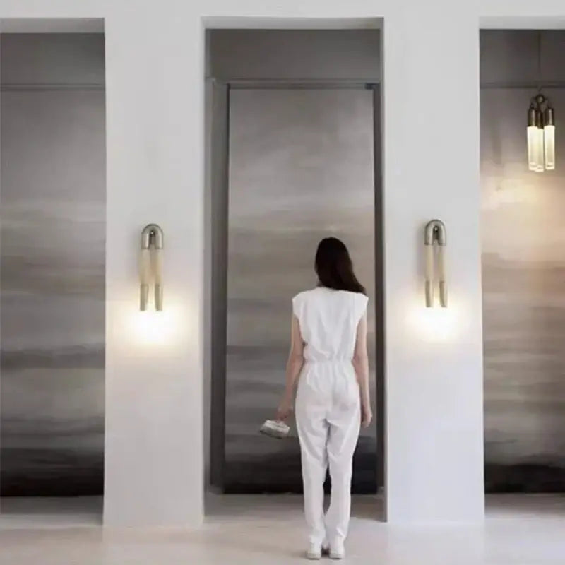 A woman in a white jumpsuit walks toward an elevator near a modern gold and black decorbites horsehair wall lamp bedroom light fixture