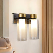Modern gold and black wall lamp featuring dual clear glass cylinders with gold-toned caps on a matte black bulb base bracket