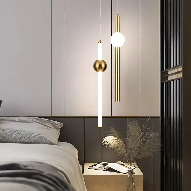Modern gold geometric line frame pendant lights with cylindrical and spherical glass globes hang above a bedside table