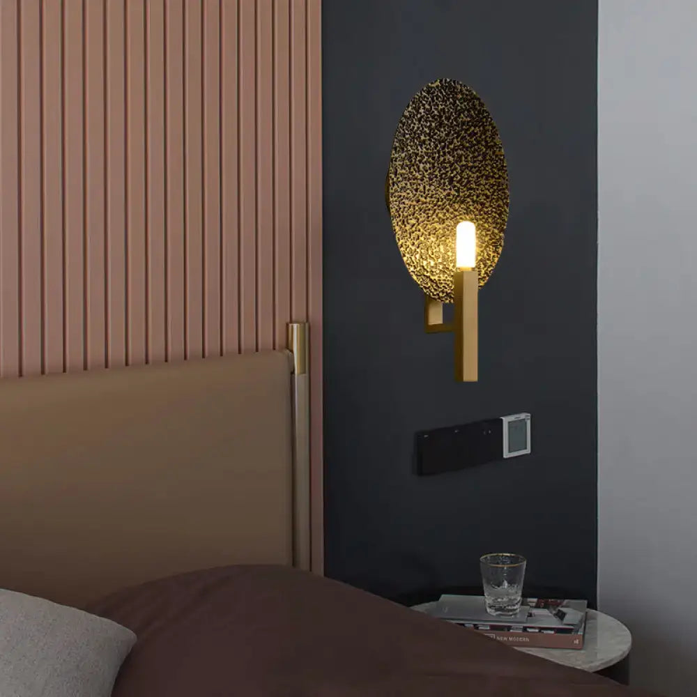Modern gold oval metal wall sconce with textured speckled glass face and sleek metal arm ideal for bedroom lighting