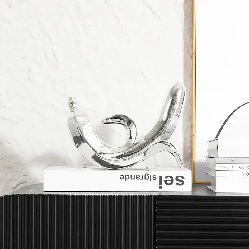 Shiny chrome abstract sculpture with flowing curved forms resting on a white book in modern golden banana figurine decor
