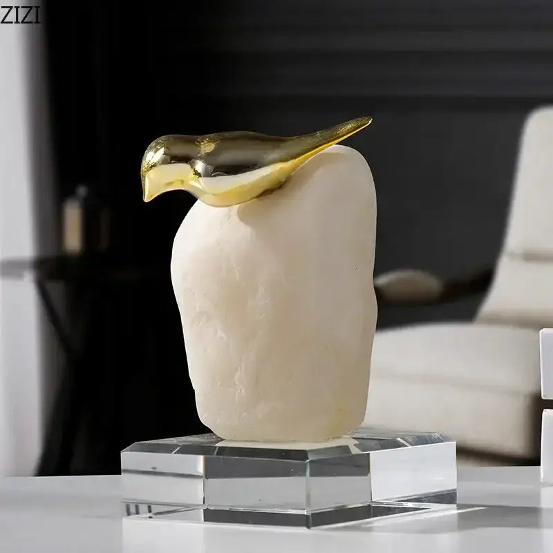 A modern golden bird statue with a polished gold beak and head on an off-white stone body and clear crystal base