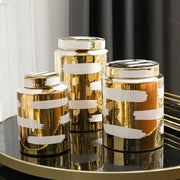 Three gold-toned ceramic striped jars with white brushstroke lids on a reflective surface for decorbites golden ceramic storage