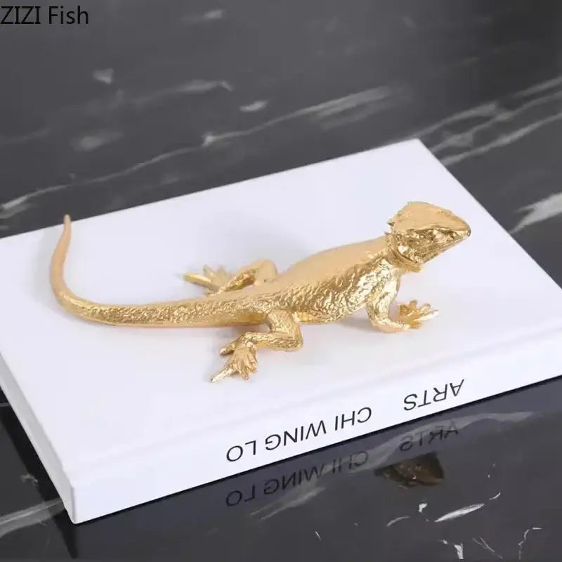 Modern golden lizard sculpture metal desk ornament with textured scales resting atop a white book