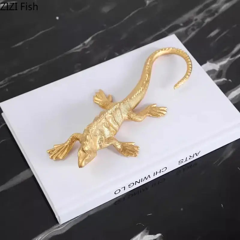 Gold-colored metal golden lizard sculpture with textured scales and curved tail resting on a white book for living room decor