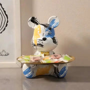 A whimsical white graffiti bear figurine with blue yellow and black patterns holds a golden tray with colorful candies