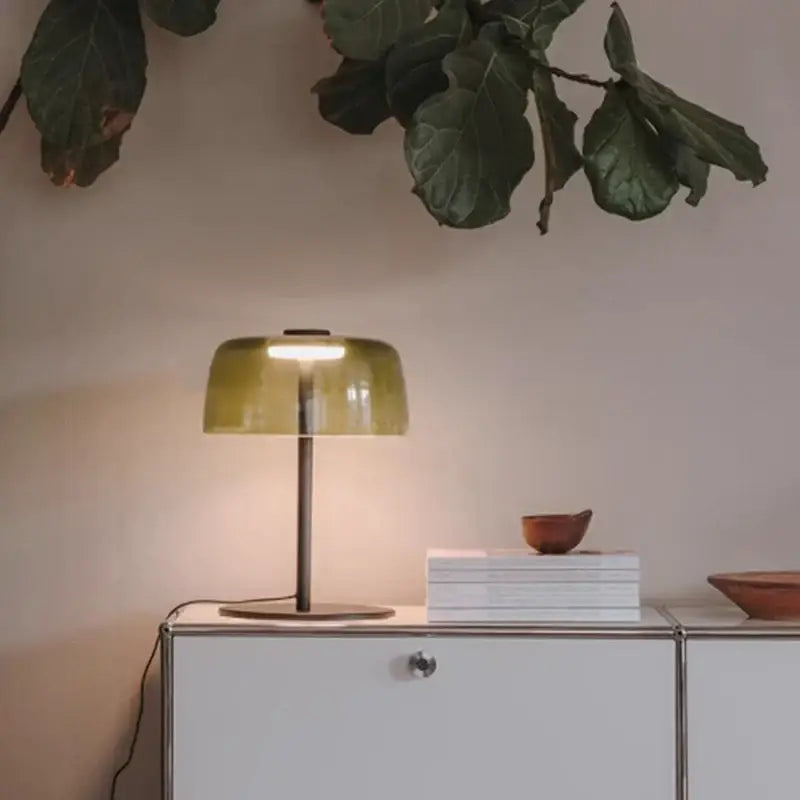 A sleek modern green glass table lamp with an olive-green translucent shade and slender metallic base for bedroom use