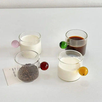 Four clear glass espresso cups with colorful marble sphere handles in pink green red and yellow with saucers