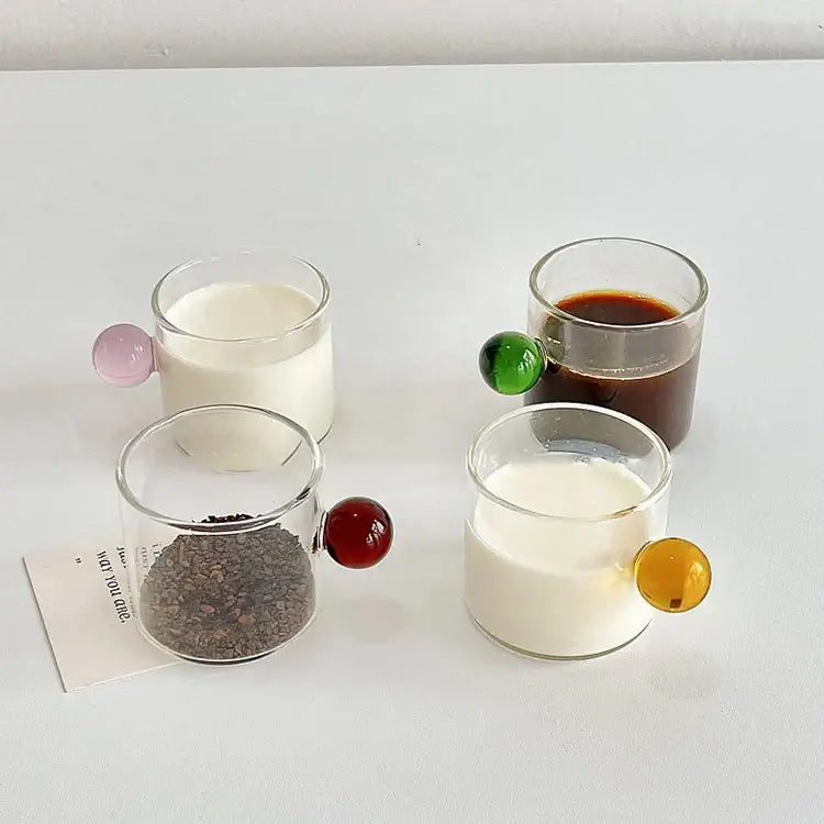 Four clear glass espresso cups with colorful marble sphere handles in pink green red and yellow with saucers