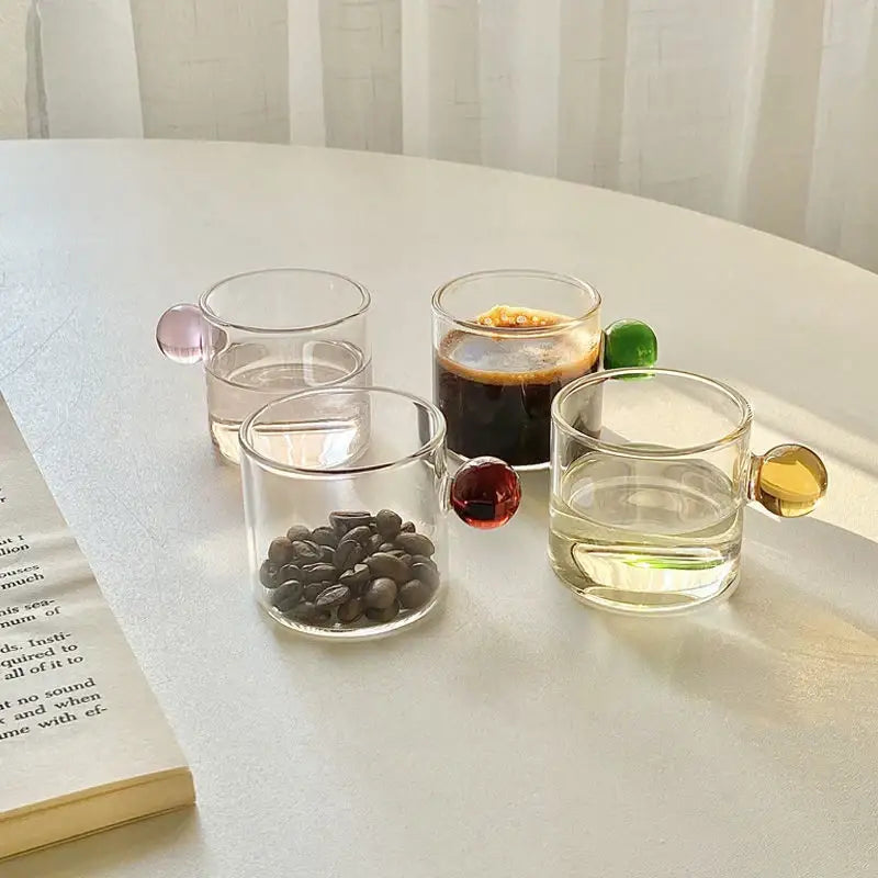 Set of four clear glass espresso cups with colorful pink red and gold spherical handles and marble saucers