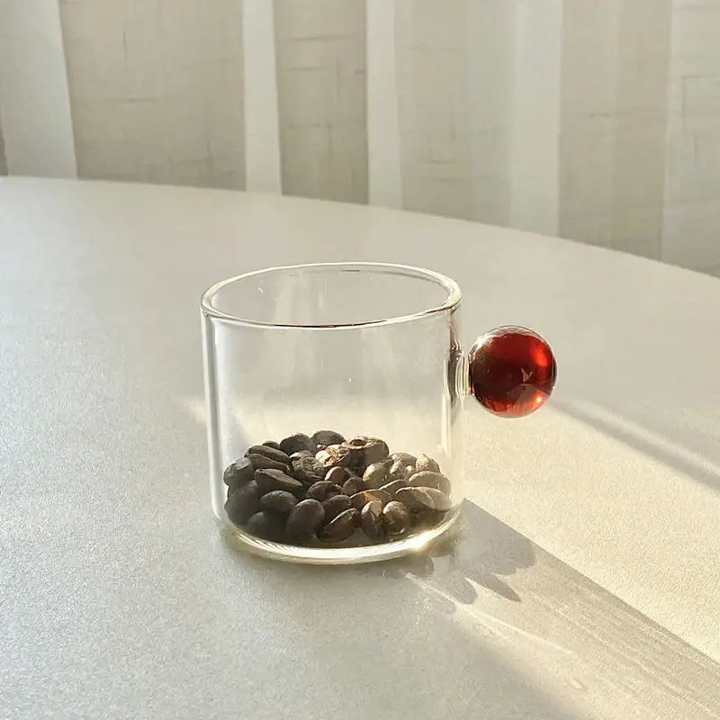 Clear glass espresso cup with a glossy red spherical handle filled with dark roasted coffee beans from modern marble sphere set