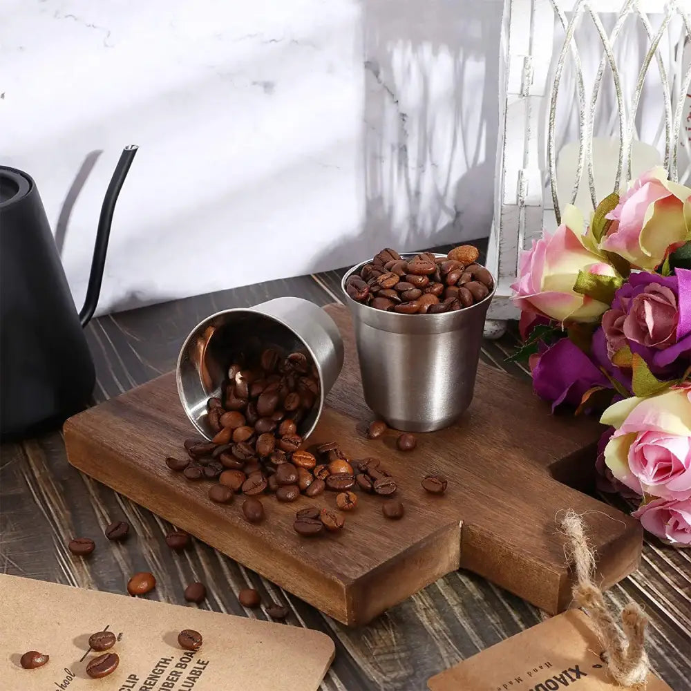 Two stainless steel modern matte double walled espresso cups with roasted beans spilling on wooden board