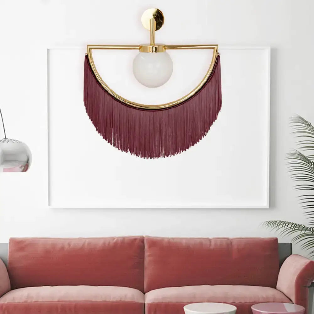 Gold-framed wall light with matte white frosted glass globe and deep burgundy tassel fringe for bedroom decor