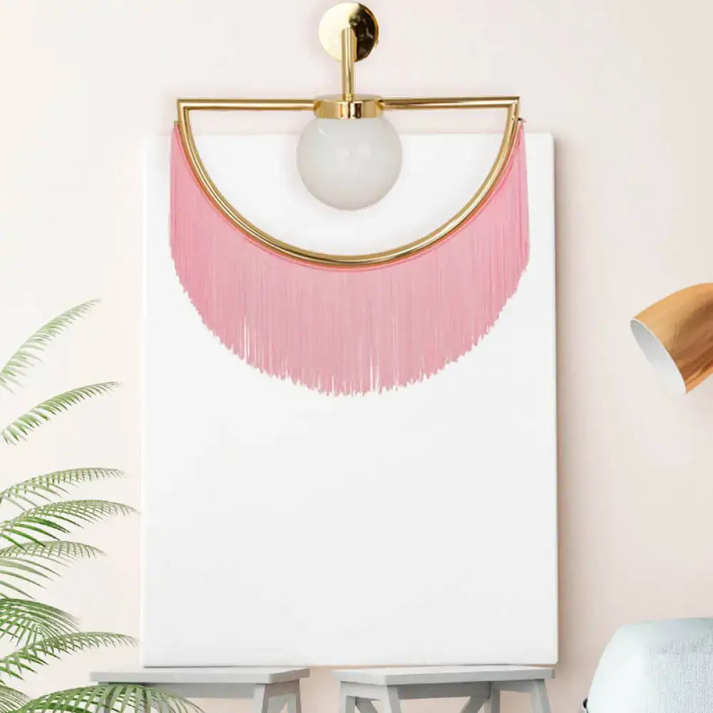 A modern matte white glass wall light with gold metal frame frosted globe bulb and cascading pink tassel fringe