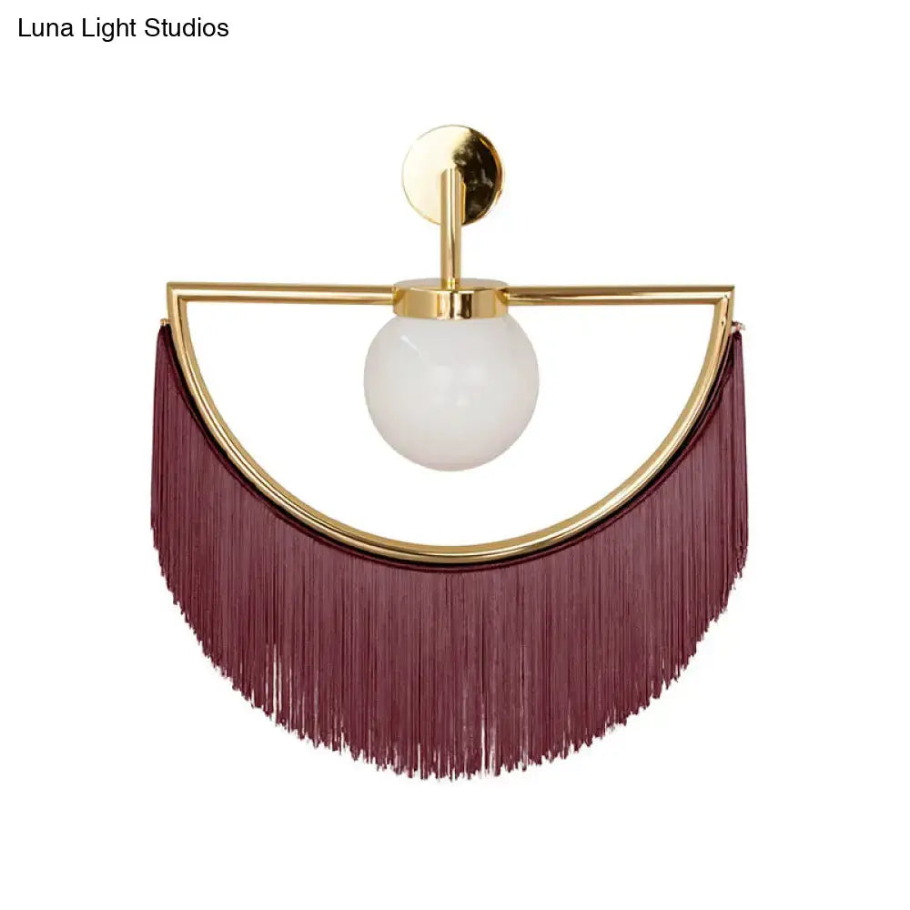 A matte white glass wall light with gold frame and maroon tassel fringe designed as a modern bedroom sconce
