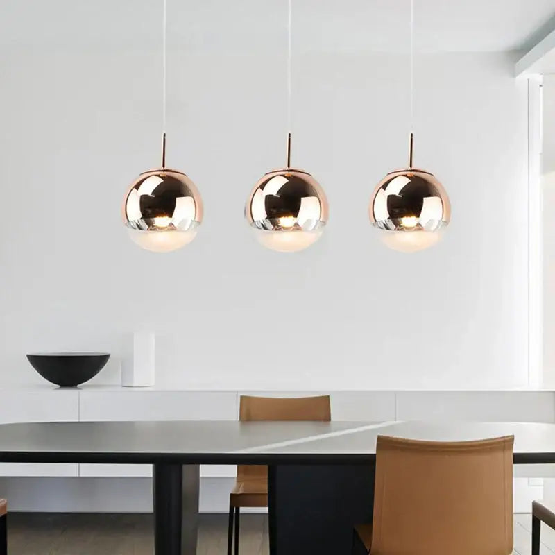 Three sleek rose gold metallic ball pendant lights with reflective surfaces hanging above a modern dining table