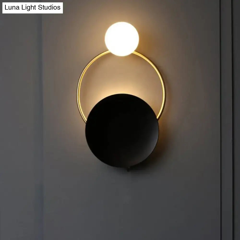 Modern minimalist black and gold wall light with a glowing white globe and matte black disc bedroom sconce