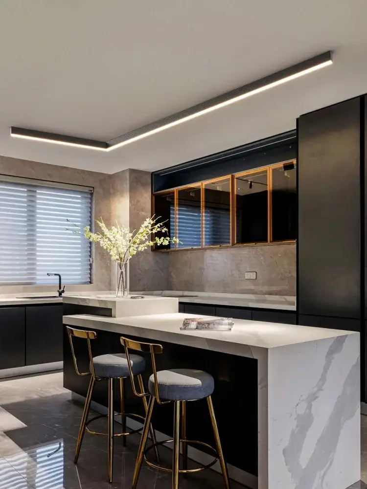 Modern minimalist linear led ceiling lamp above white marble kitchen island with black cabinetry and gold bar stools decorbites led ceiling