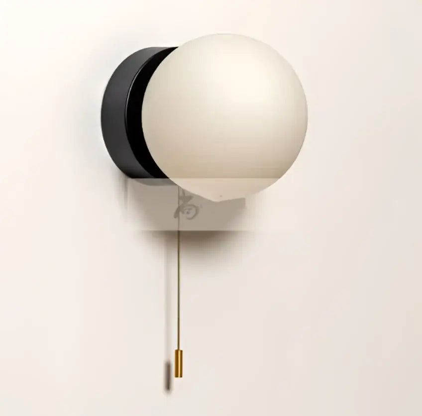 A sleek black wall light with a white frosted glass ball shade and brass pull chain for modern minimalist decor