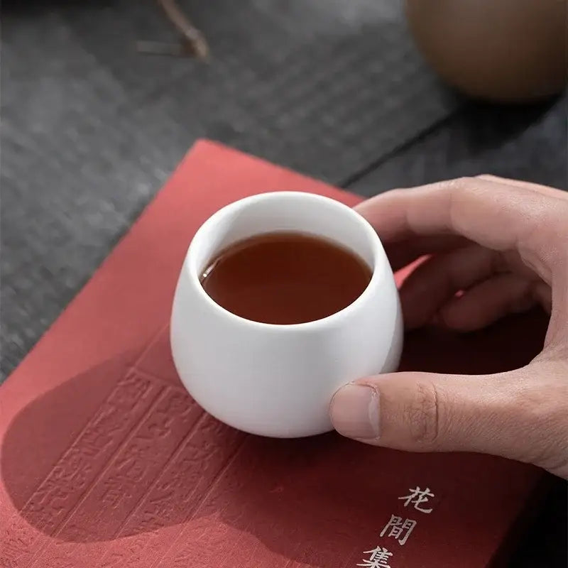 A white ceramic kungfu tea cup filled with amber tea held over a red book, part of decorbites ceramic kungfu set
