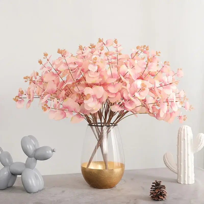 A clear glass vase with a gold-finished base holds a modern pink eucalyptus flower arrangement for living room decor