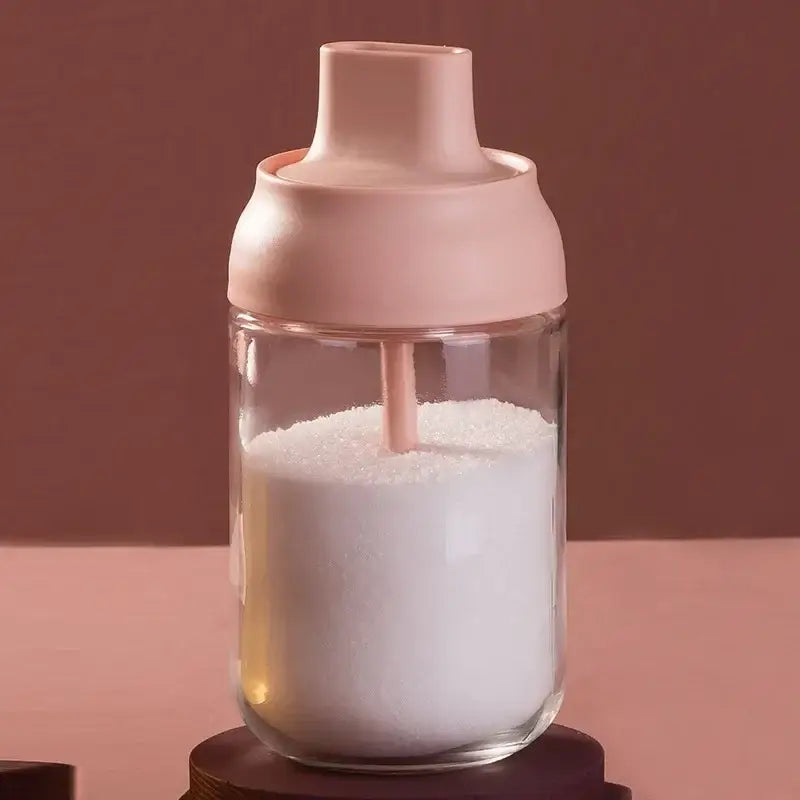 A clear glass seasoning bottle with a soft pink lid and straw filled with white granulated sugar for kitchen storage
