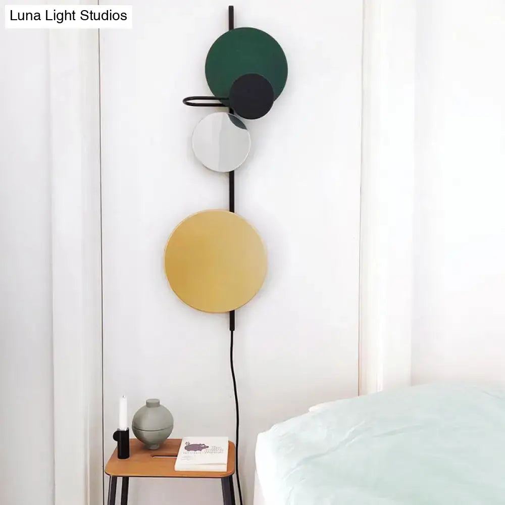 Modern geometric wall sconce with stacked circular discs in matte green, black, and gold-toned finishes mounted on a sleek black metal frame.