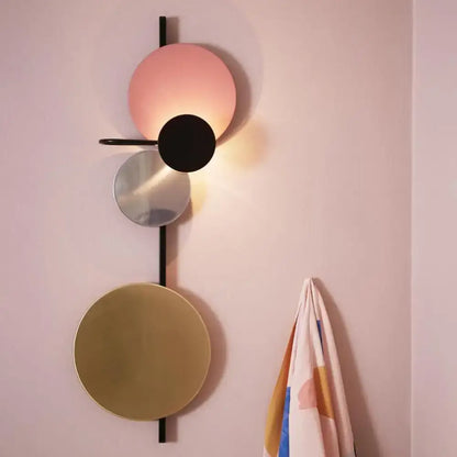 Modern geometric wall sconce featuring stacked circular discs in matte pink, black, mirrored silver, and brushed gold tones mounted on a sleek black vertical rod.