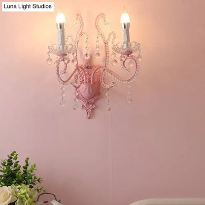 Modern pink metallic scroll arm wall lamp with crystal droplets and two candle-style bulbs for bedroom sconce lighting