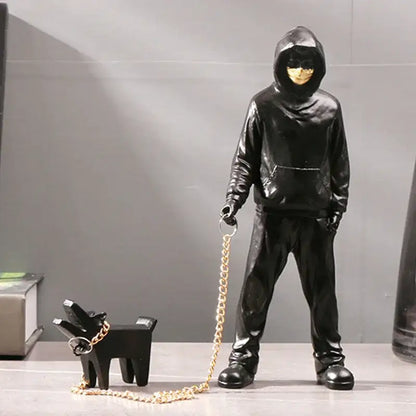Black glossy hooded figure with gold face and chain leash standing beside small black decorbites dog sculpture