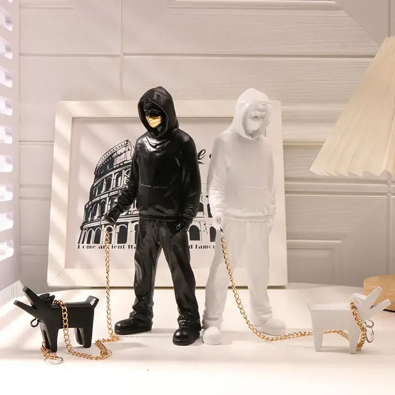 Black glossy resin hooded figure with gold mask and chain leash beside translucent white decorbites dog sculpture