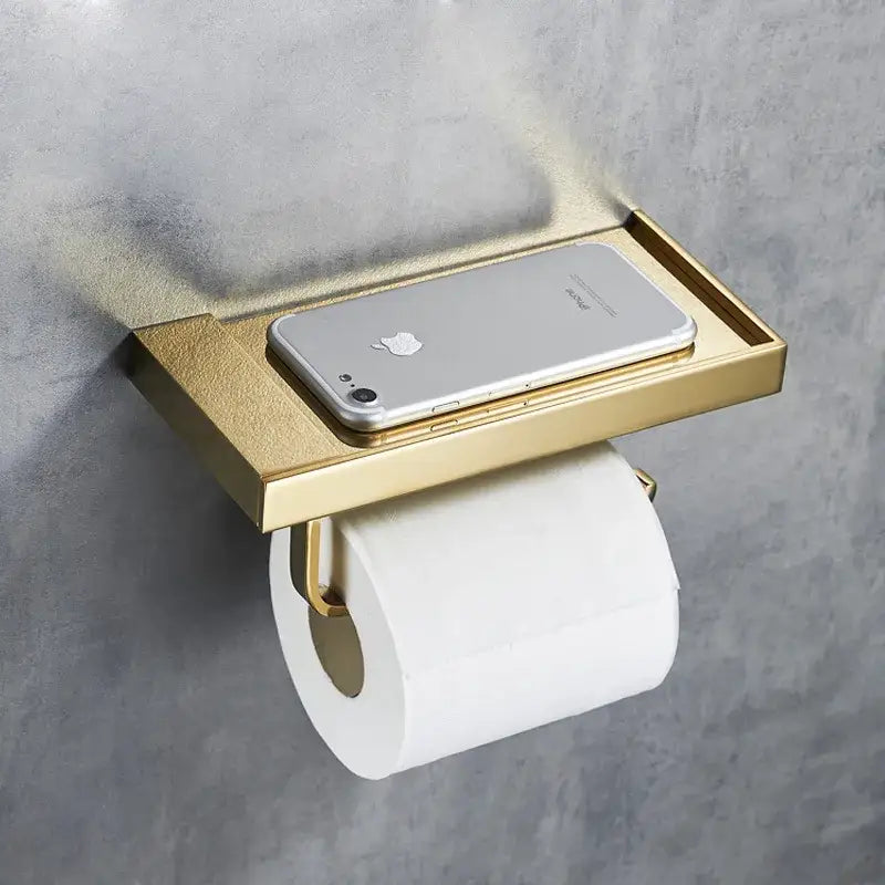 Modern rectangular gold-toned metal toilet paper holder with integrated tray holding a silver smartphone for bathroom use
