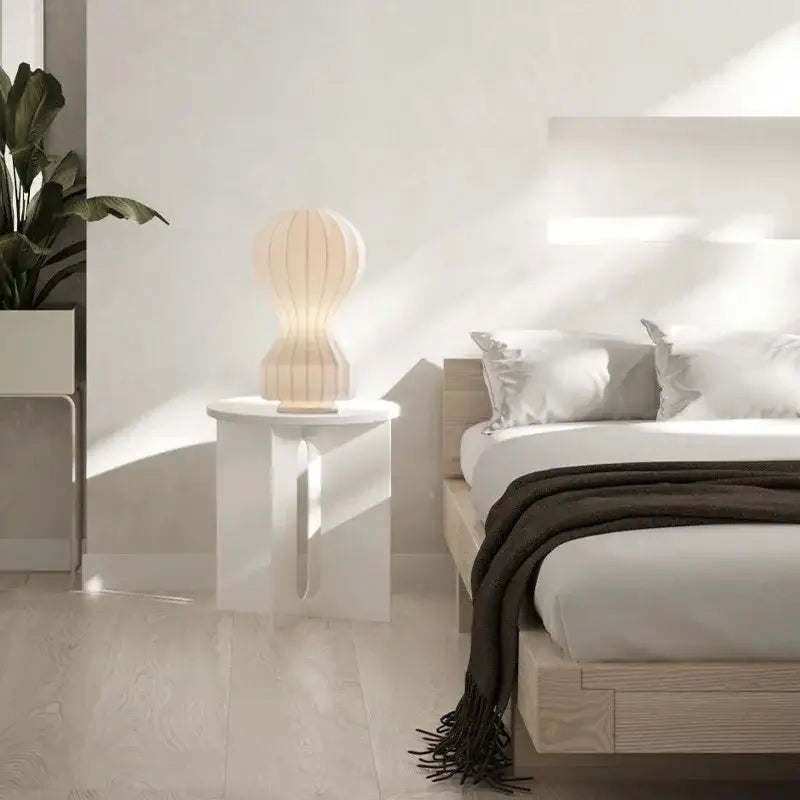 A softly glowing cream-colored silk table lamp with a ribbed ceramic design on a white minimalist side table