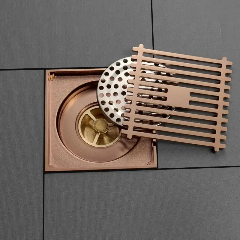 Modern rose gold brushed copper-toned square floor drain with slatted grate and circular perforated cover for bathroom use